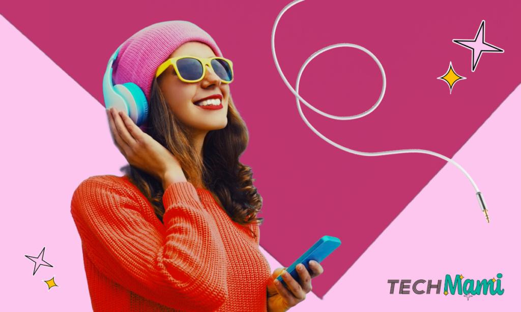 Eine junge Frau in einem leuchtend roten Pullover und einer rosa Mütze genießt Musik über große, bunte Kopfhörer. Sie trägt gelbe Sonnenbrillen und hält ein Smartphone in der Hand. Der Hintergrund ist lebhaft rosa gestaltet, und das TechMami-Logo mit dekorativen Sternen schmückt die obere Ecke.